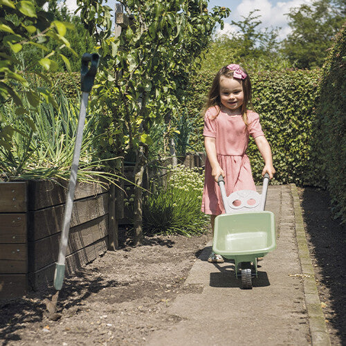 Een jong meisje duwt de dantoy speelgoed kruiwagen over een pad in de tuin.