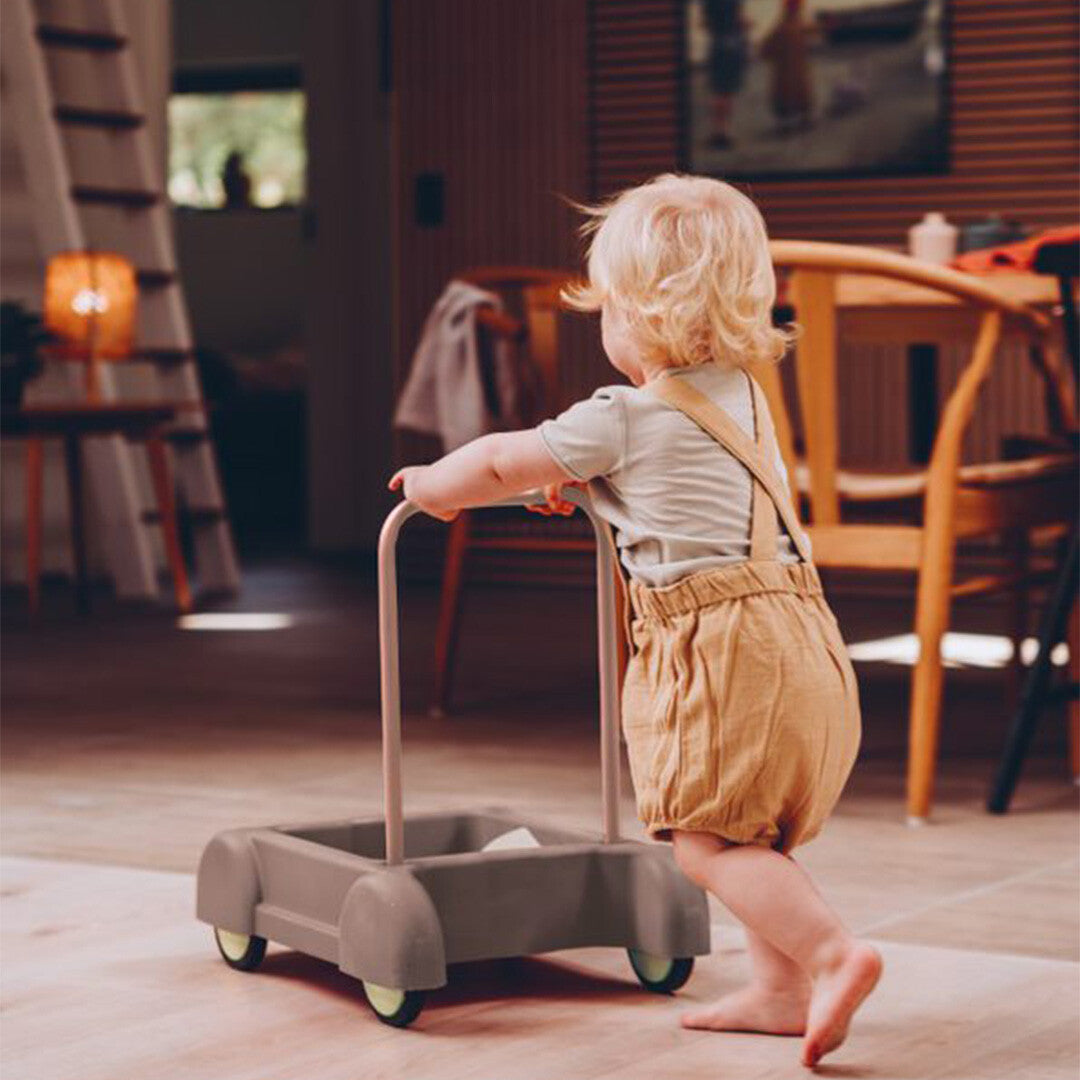 Een baby met blonde haren duwt de dantoy grijze loopwagen over een houten vloer in huis.