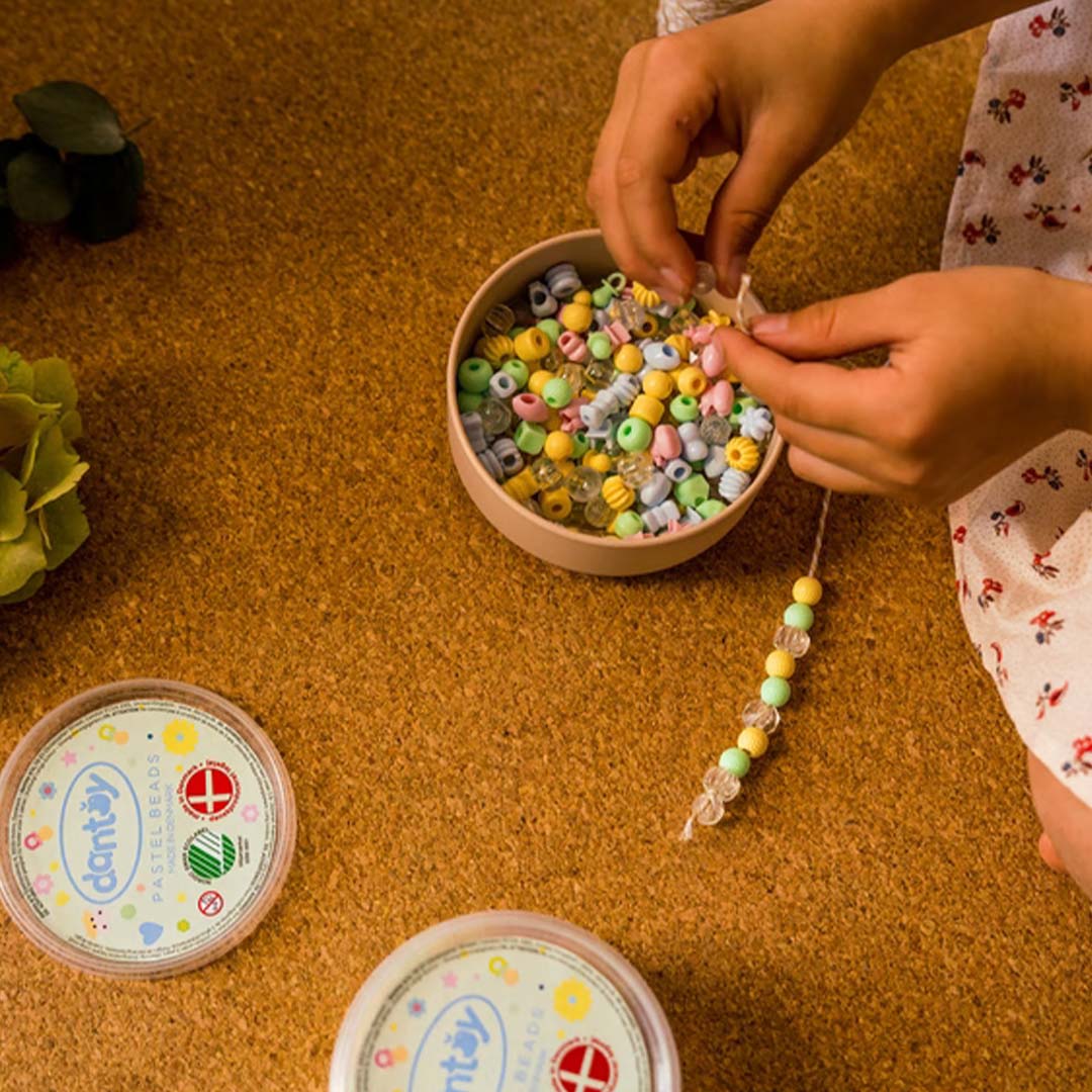Kinderhanden rijgen pastelkleurige kralen van de dantoy kralenset aan een touwtje, met twee verpakkingen op de achtergrond.