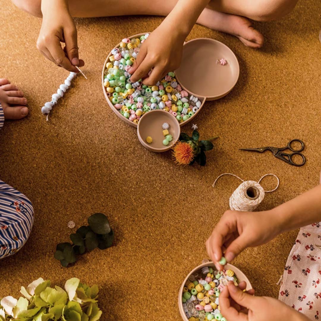 Meerdere kinderhanden rijgen pastelkleurige kralen van de kralenset aan een touwtje op een kurkvloer.