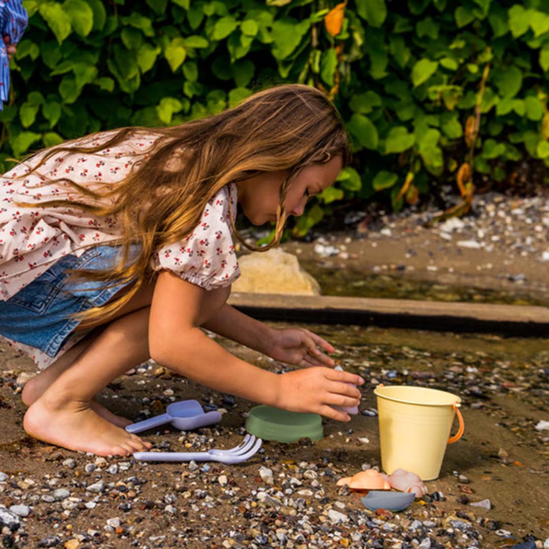 Een kind speelt met de dantoy pastel zandset op een kiezelstrand met groen in de achtergrond.