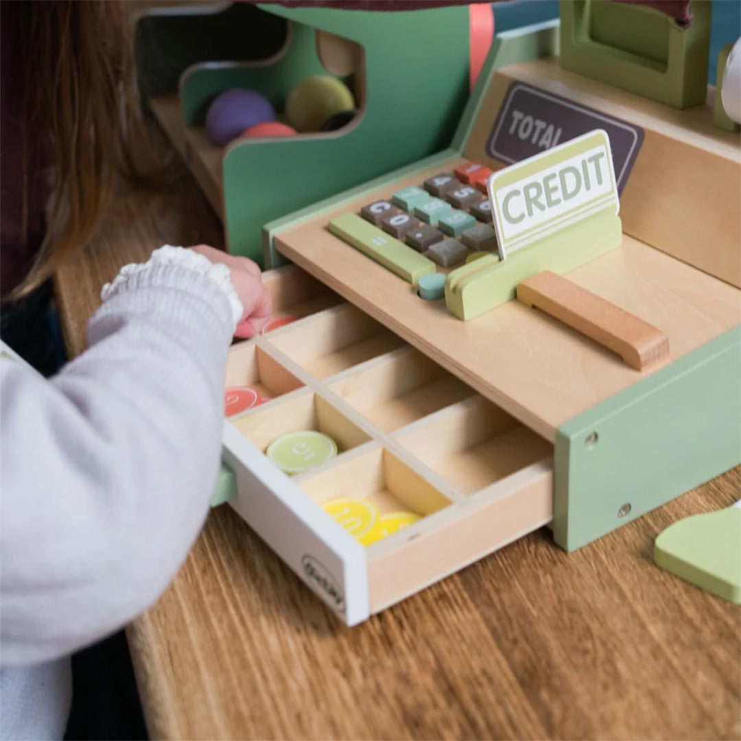 Een kinderhand pakt een houten munt uit de open lade van de dantoy houten kassa op een houten tafel.