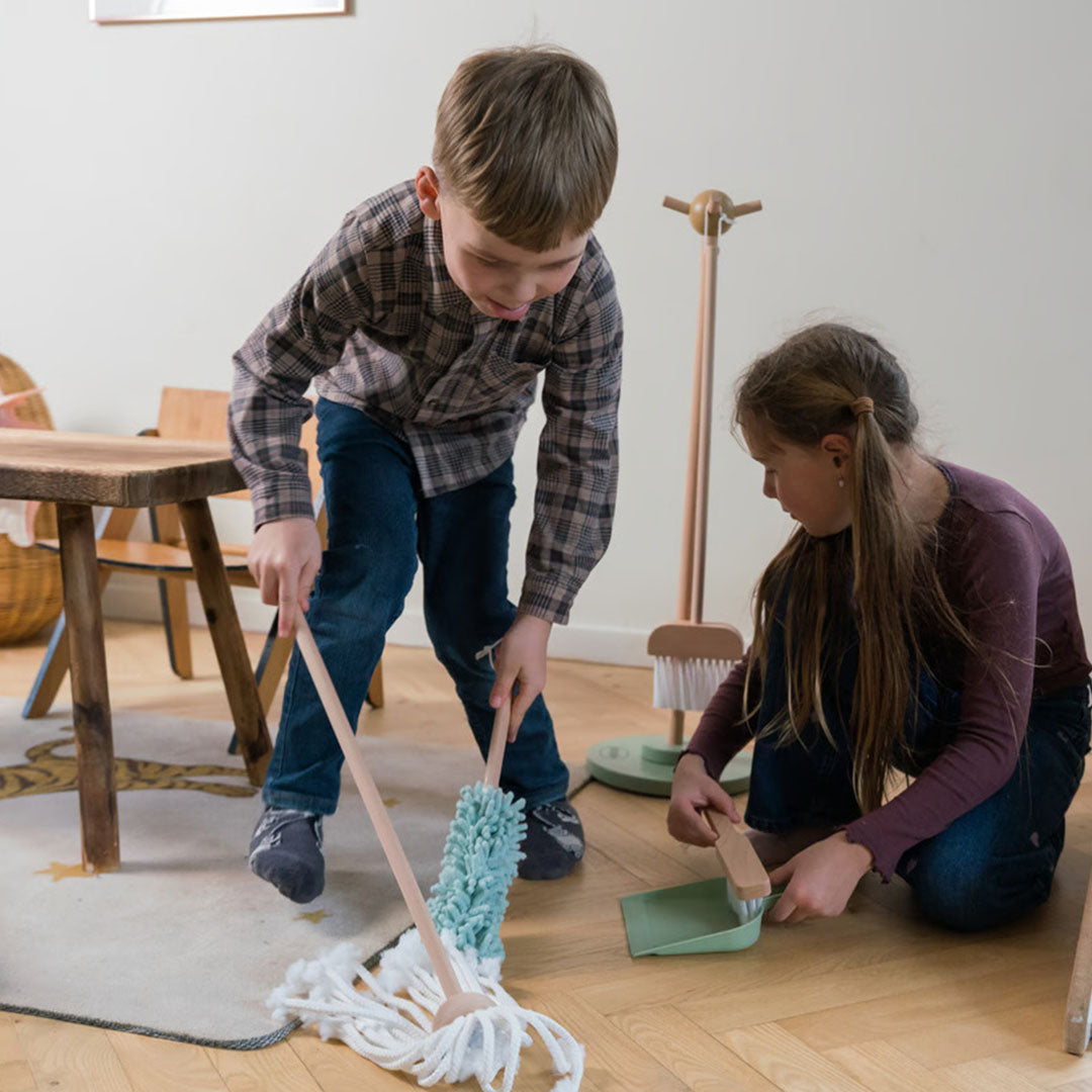 Twee kinderen spelen met de houten speelgoed schoonmaakset, waarbij een jongen dweilt en een meisje veegt.