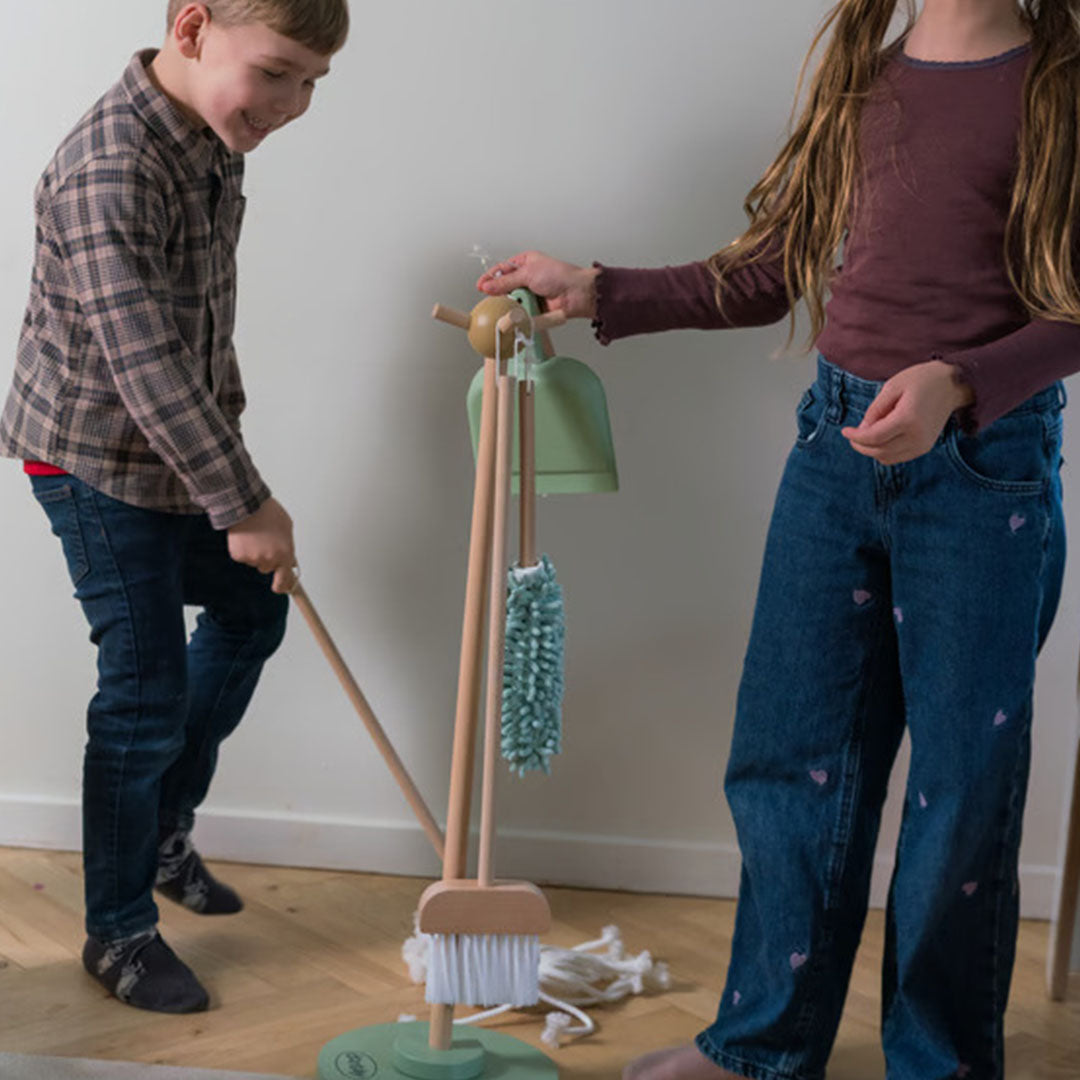 Twee kinderen spelen met de houten speelgoed schoonmaakset, waarbij een jongen veegt en een meisje een plumeau pakt.