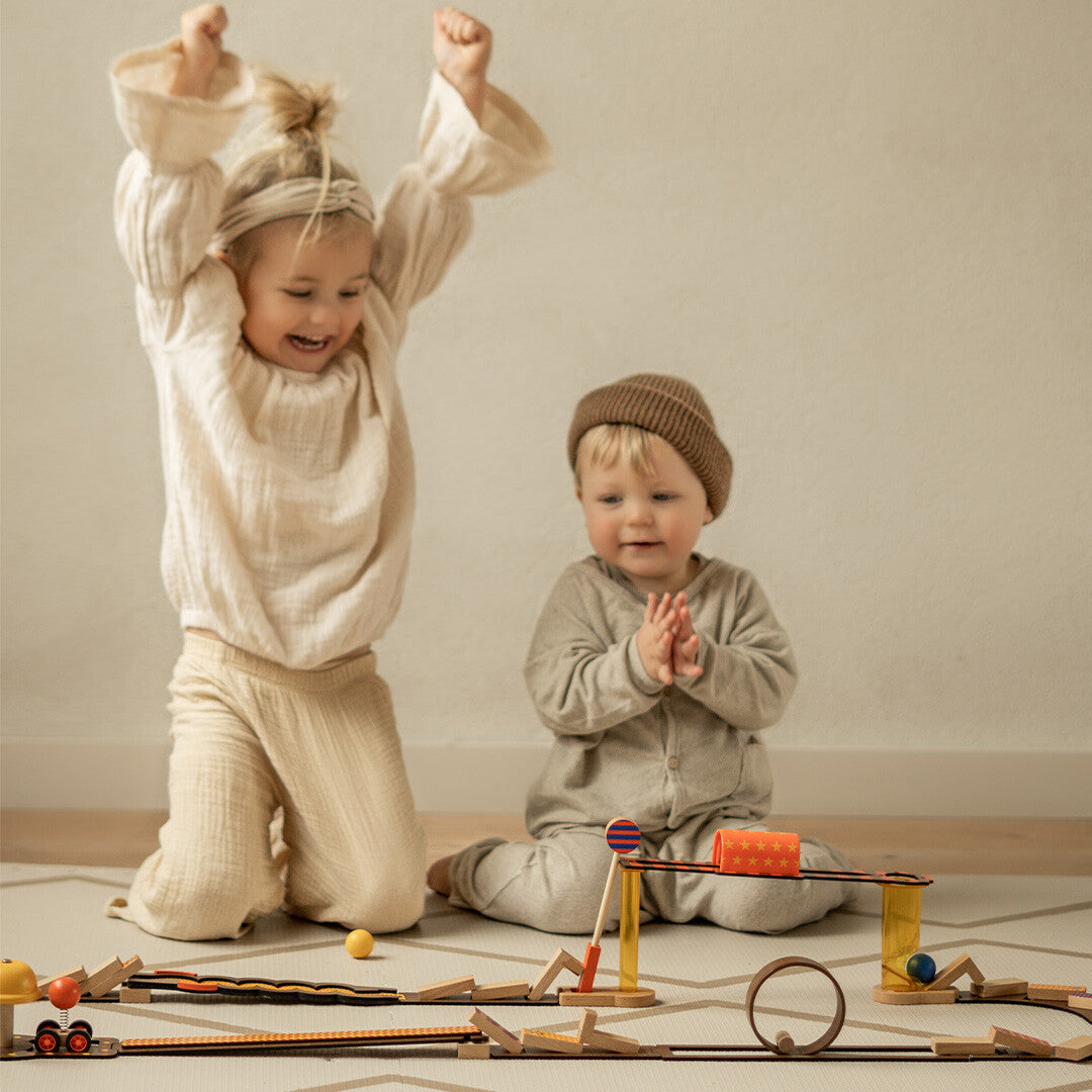 Twee lachende kinderen spelen met het Djeco Zig & Go Junior kettingreactiespel op de vloer.