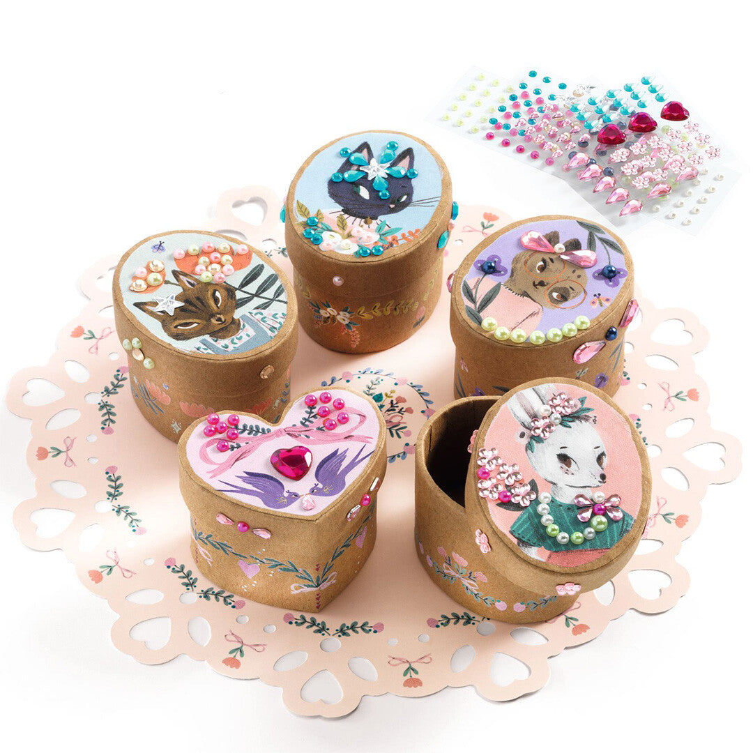 Vijf versierde Djeco DIY opbergdoosjes met dieren en bloemen, op een roze onderlegger met hartjes.
