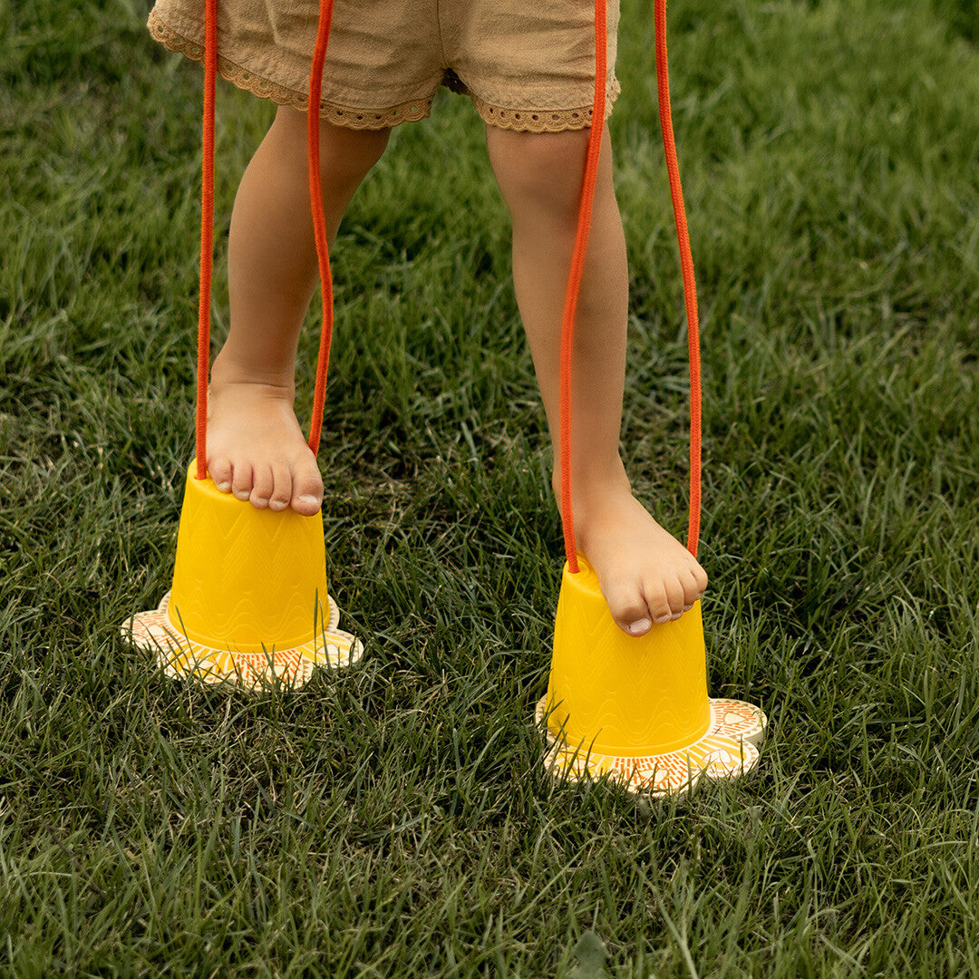 Close-up van blote kindervoeten op gele loopklossen met oranje touwen in het gras.