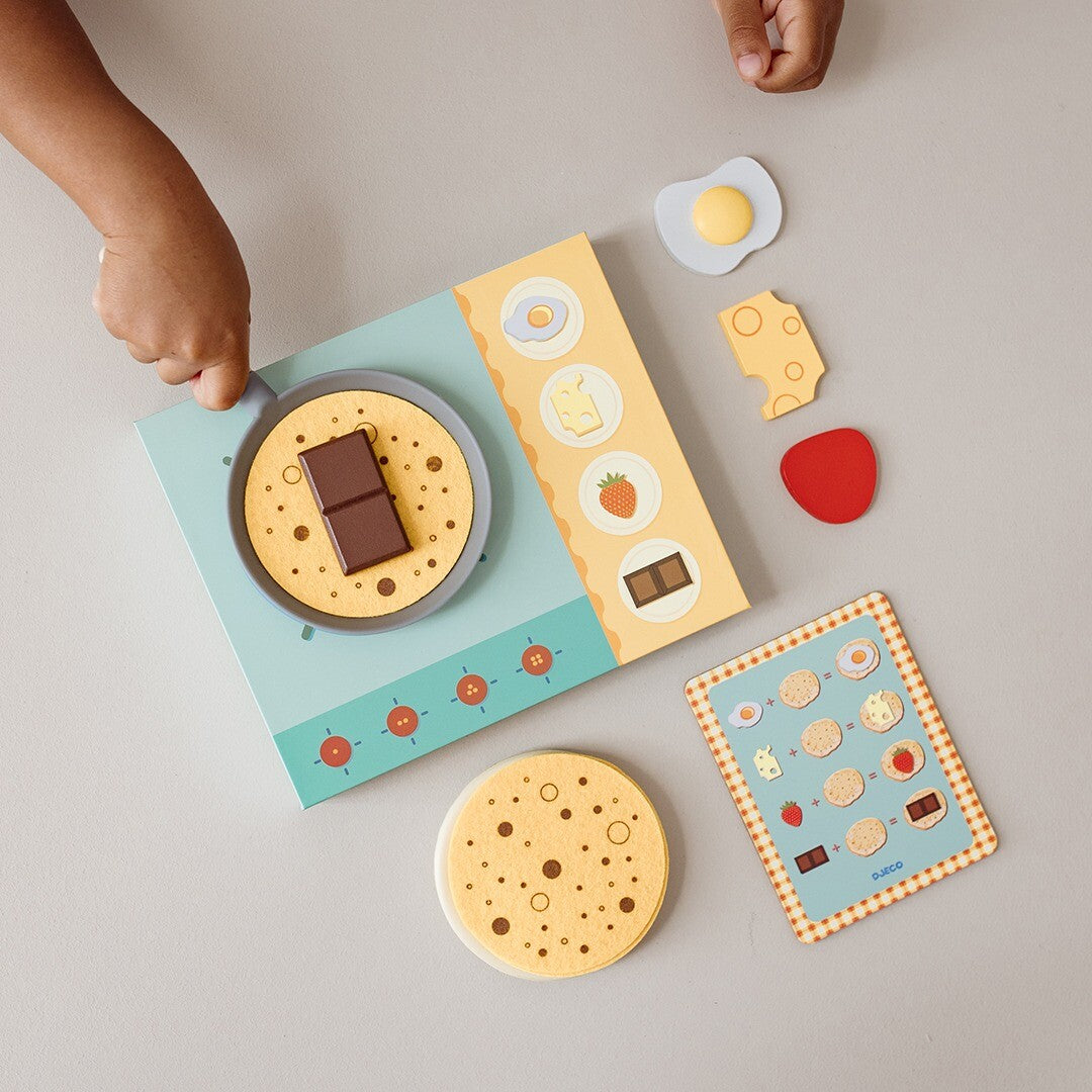 Bovenaanzicht van kinderhanden die spelen met de Djeco pannenkoeken keukenspeelset, met een pan op het fornuis en losse toppings.