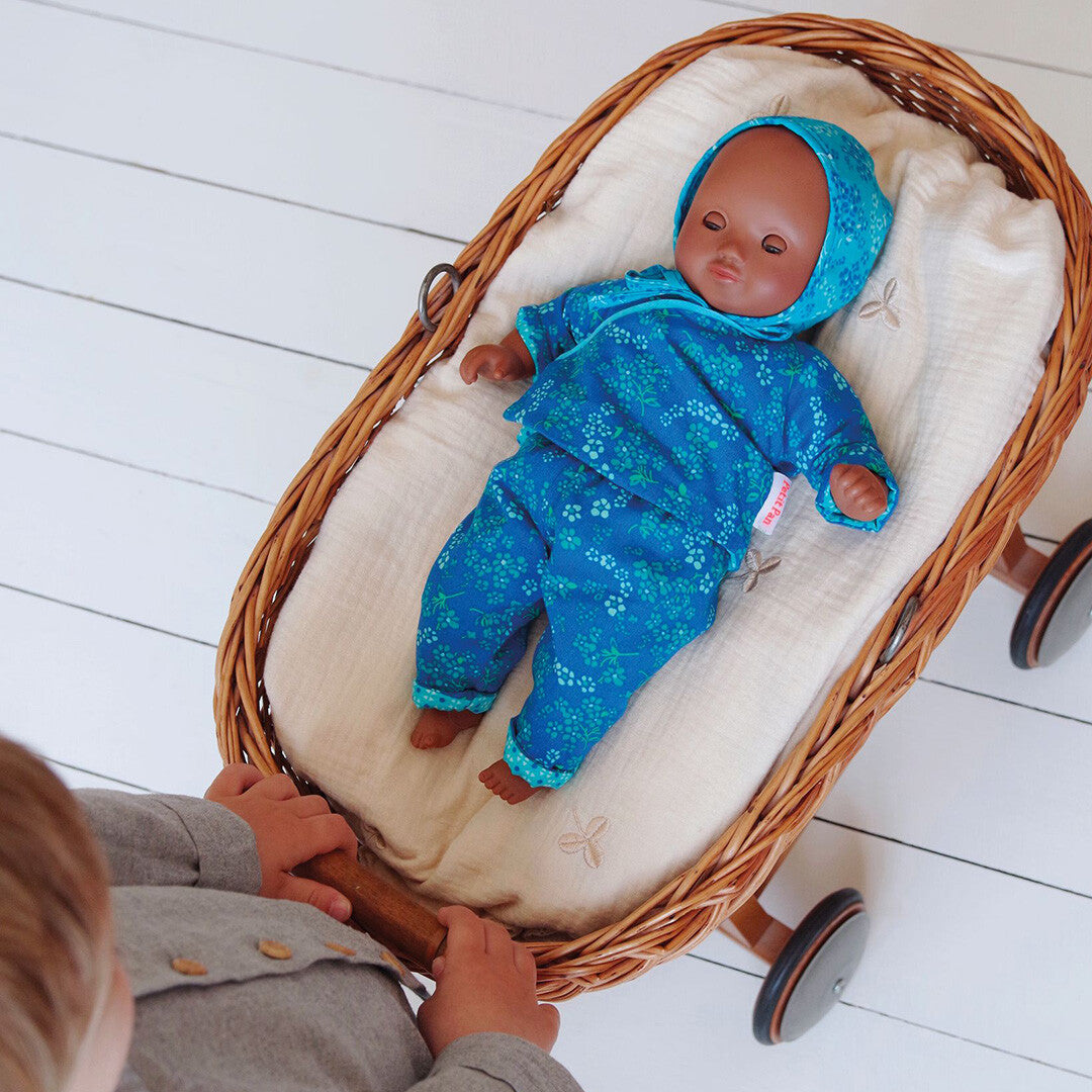 Een pop met de Djeco blauw gebloemde poppenkledingset ligt in een rieten poppenwagen die door een kind wordt geduwd.