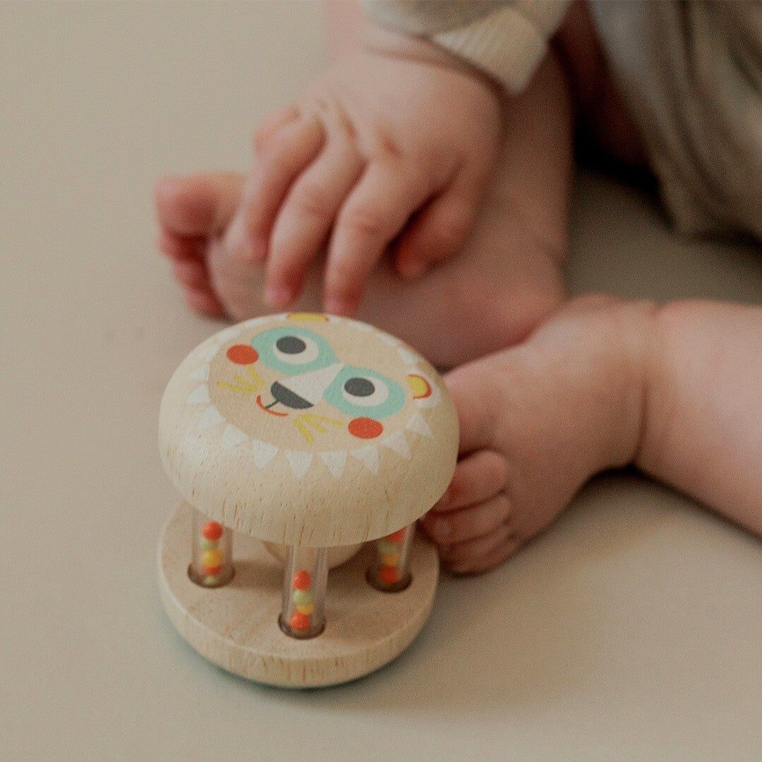 Djeco houten rammelaar met leeuwengezicht op een lichte ondergrond, met babyvoetjes en -handjes op de achtergrond.