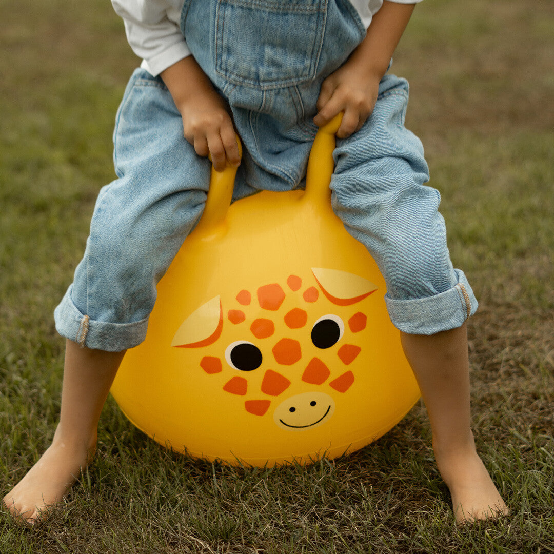 Een kind zit op een gele skippybal met girafgezicht en houdt de handvatten vast, met blote voeten in het gras.