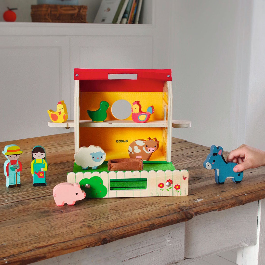 Een kind speelt met de Djeco houten speelboerderij op een houten tafel, met een hand die het ezeltje vasthoudt.