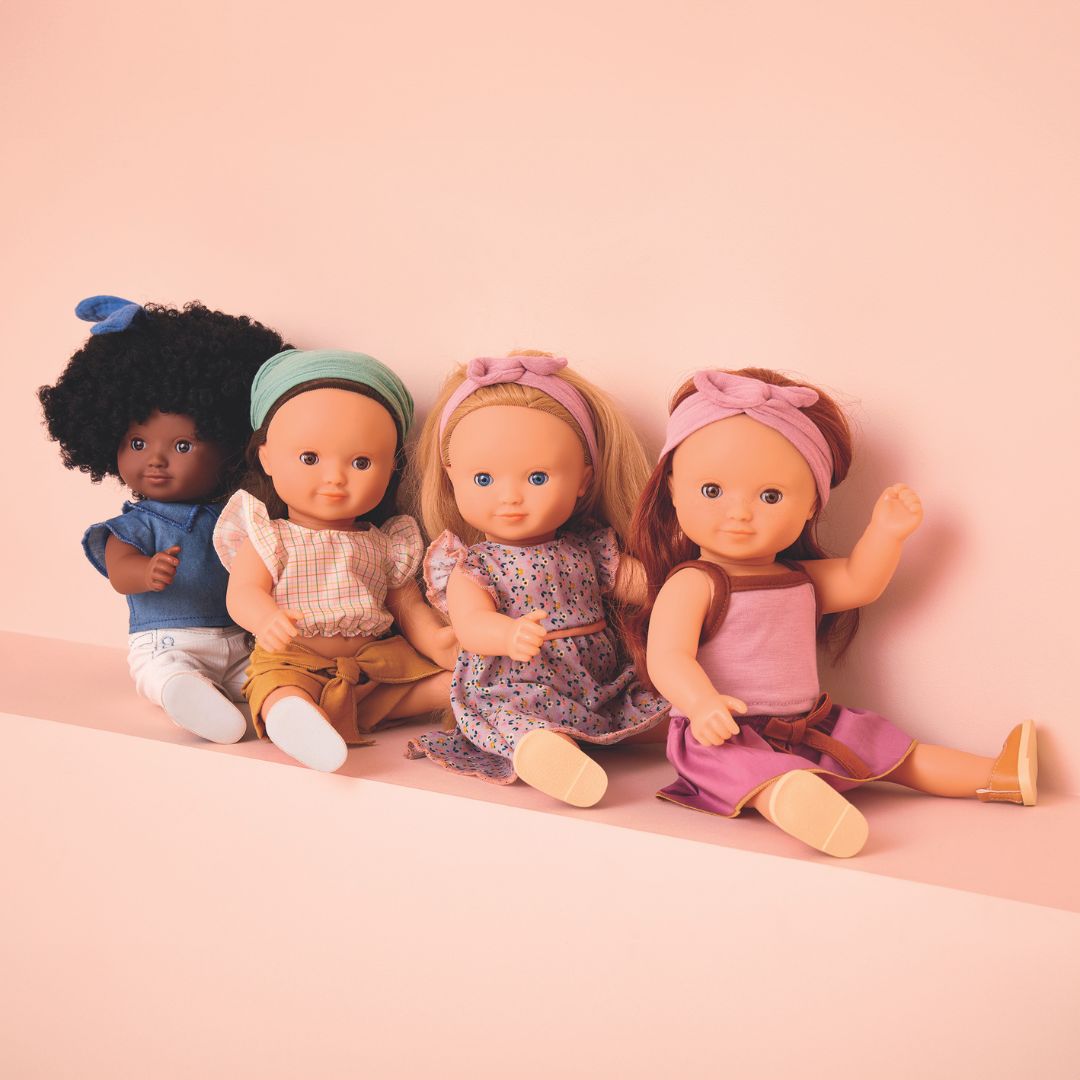 Vier Djeco modepoppen met verschillende huidskleuren en outfits zitten naast elkaar op een roze ondergrond.