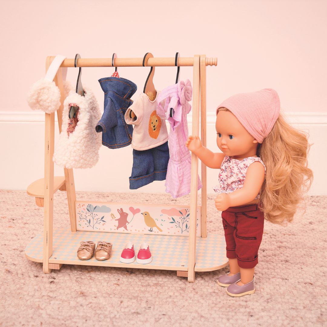 Djeco stapop Lovely Marie staat naast een houten kledingrek met diverse poppenkleding.