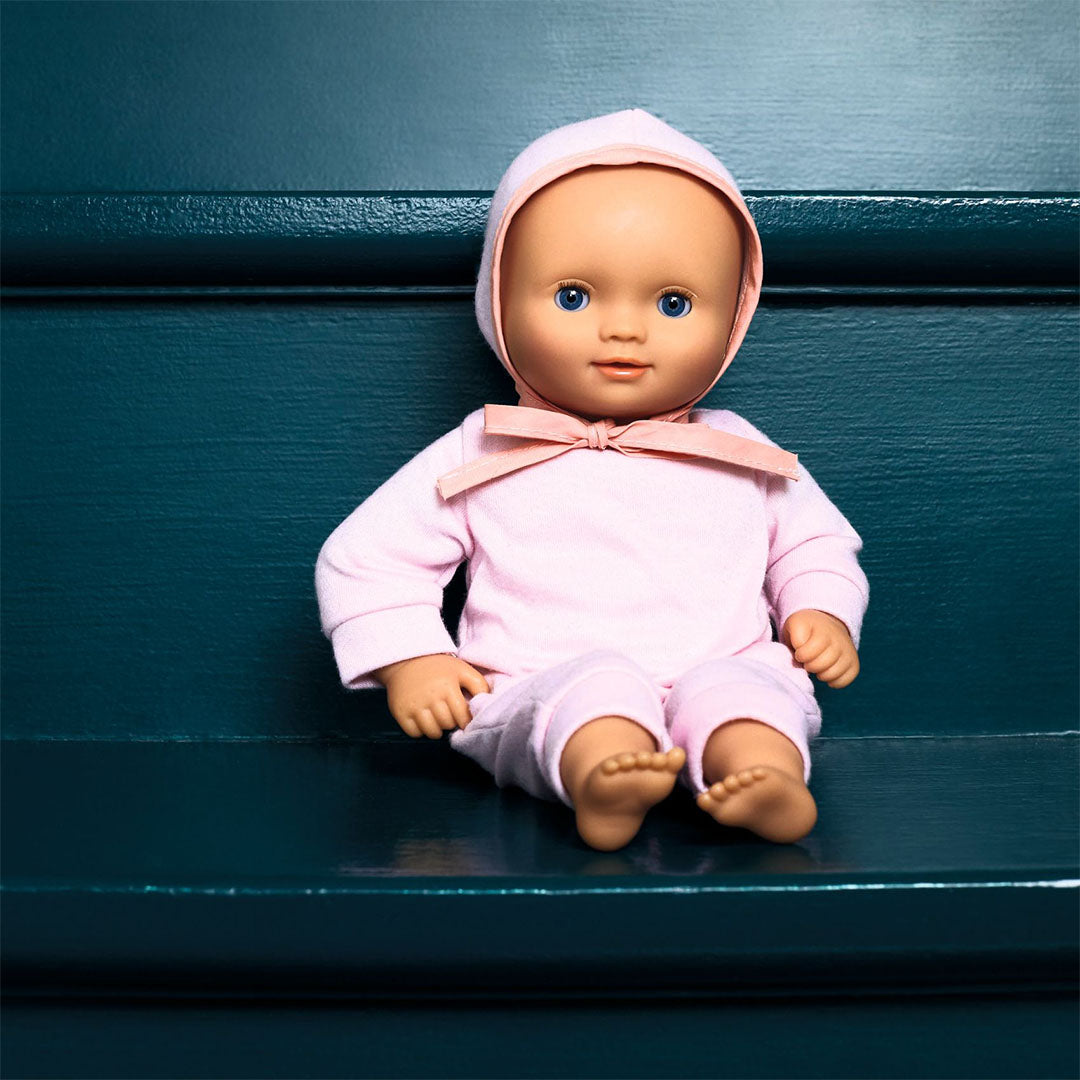 Djeco babypop Lilas Rose in roze romper en mutsje, zittend op een donkergroene ondergrond.