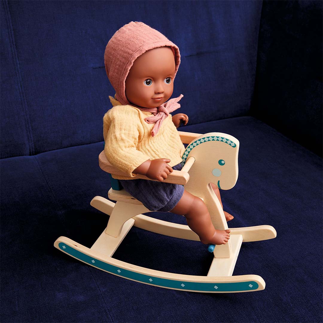 Djeco babypop Mimosa zittend op een houten hobbelpaard op een blauwe bank.
