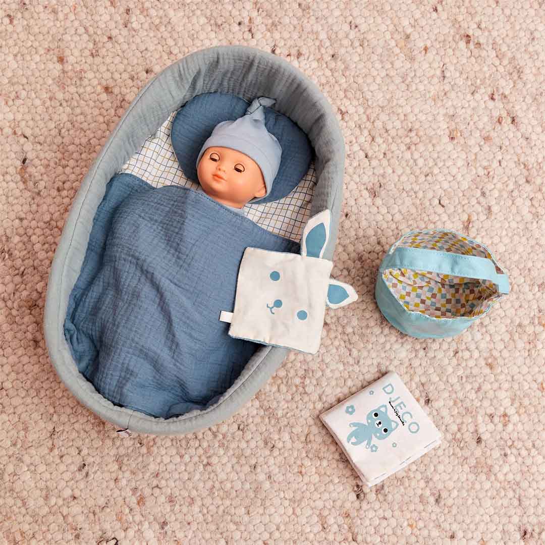 Djeco babypop Pomea in een draagmandje met blauwe deken en muts, naast een konijnenknuffeldoekje en een mandje.