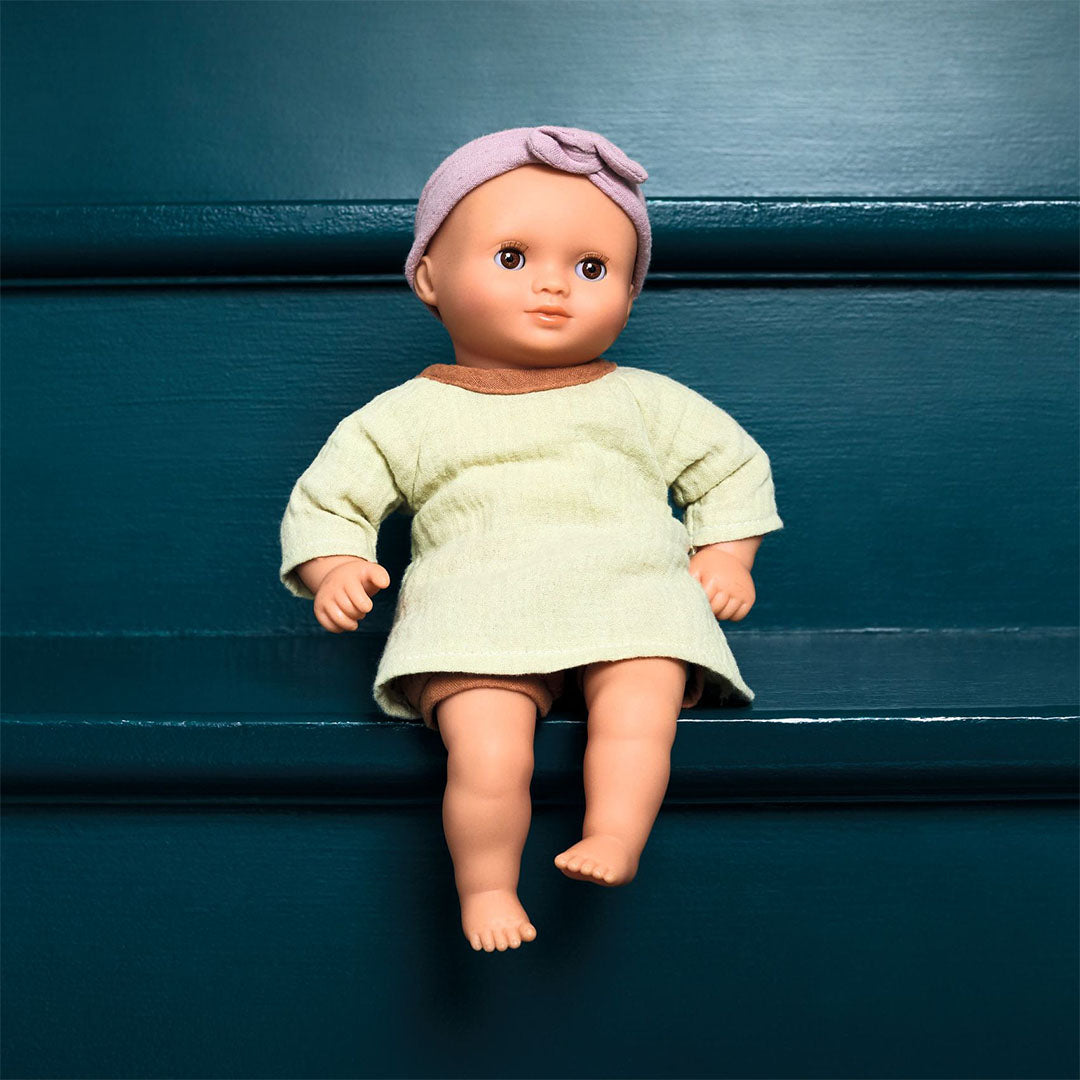 Djeco babypop Pomea met lichtgroen jurkje en roze hoofdband, zittend op een donkerblauwe ondergrond.