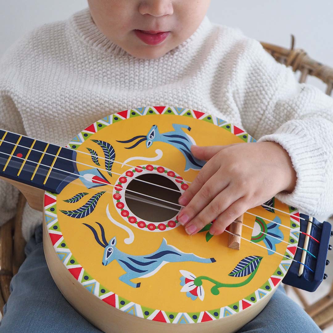 Close-up van een kinderhand die de snaren van de Djeco houten speelgoedbanjo met hertenpatroon bespeelt.