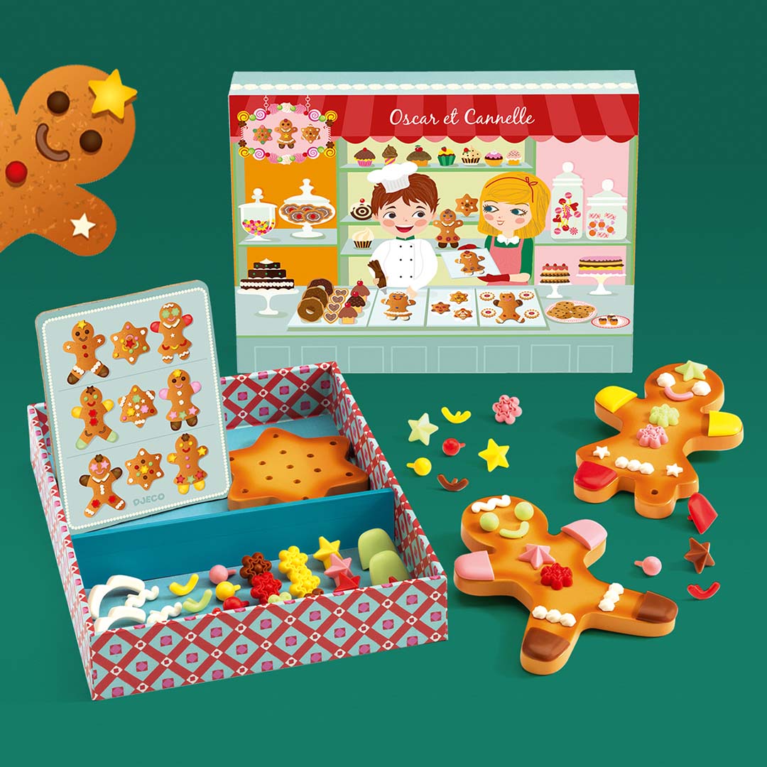 Djeco speelgoedset met twee versierde gingerbread koekjes, een open doos met onversierde koekjes en decoraties, en een bakkerij achtergrondkaart op een groene ondergrond.
