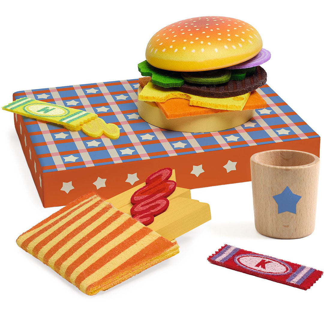 Een close-up van de Djeco speelgoed hamburger, frietjes met ketchup en een houten beker op een geruite ondergrond.
