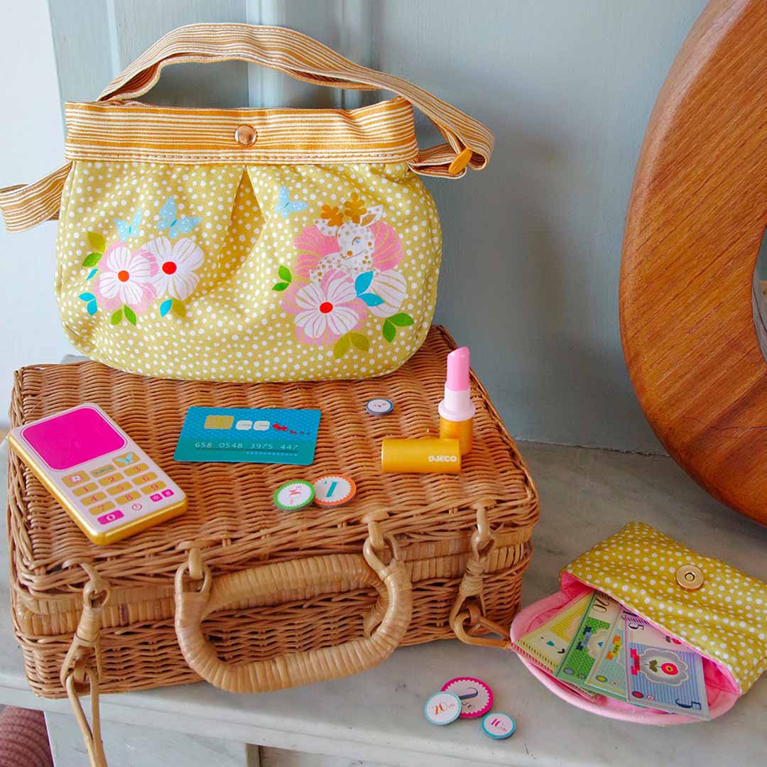 Djeco geel speelhandtasje met bloemen en bijpassende accessoires op een rieten koffer.