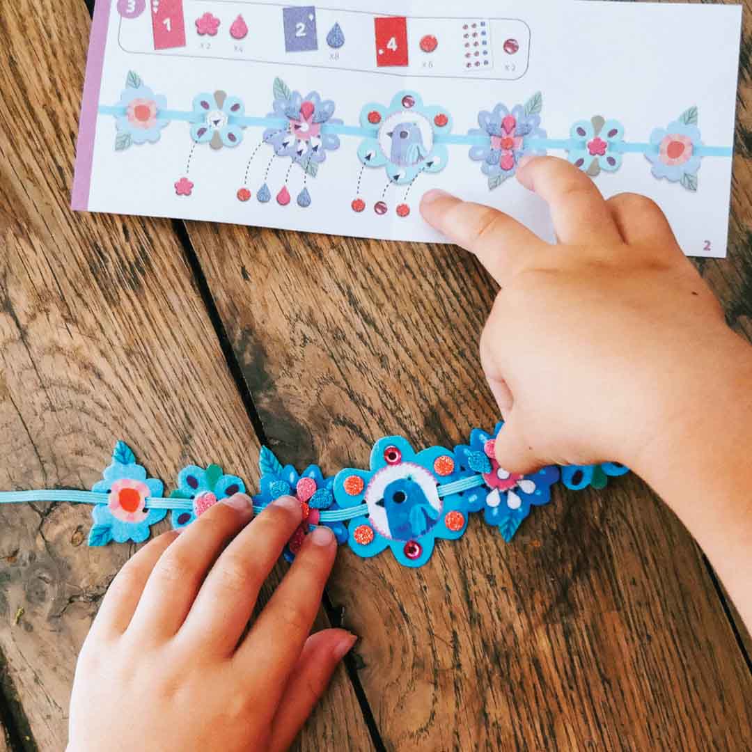 Kinderhanden knutselen een vilten armbandje met bloemen en een vogel op een houten tafel.
