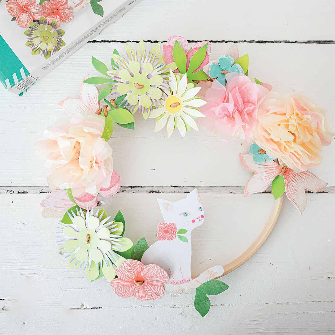 Een afgewerkte bloemenkrans met diverse papieren bloemen en een witte kat op een houten ring.