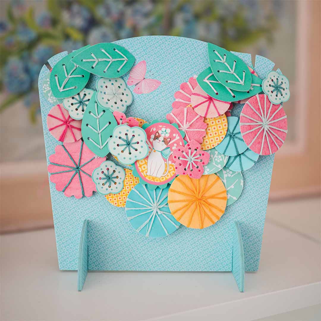 De gemaakte bloemenketting van de knutselset Flora, gepresenteerd op een lichtblauwe standaard.