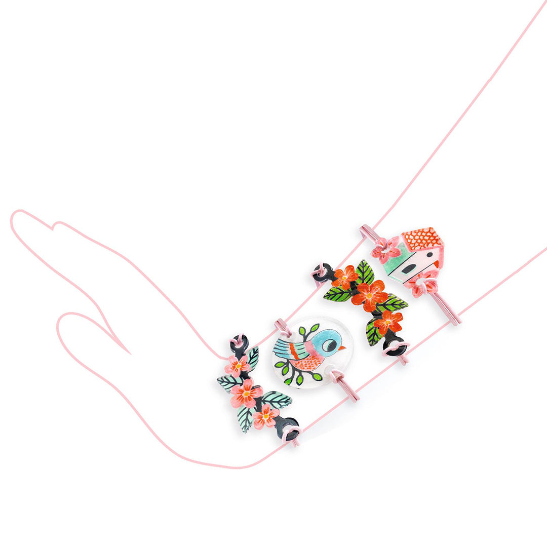 Drie armbandjes uit de Djeco sieradenset met bedels van een vogel, bloemen en een huisje, gedragen om een getekende hand.