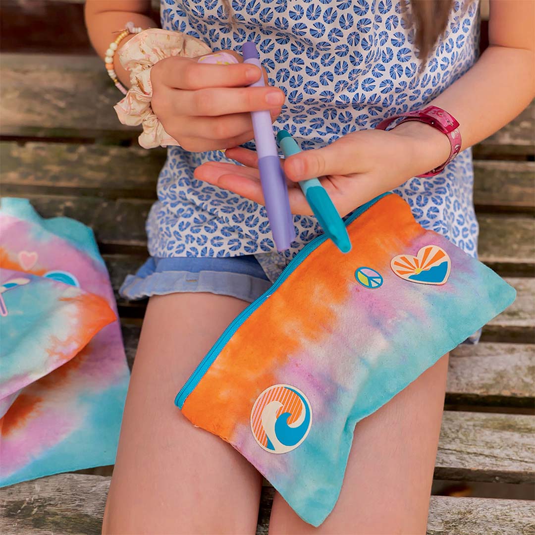 Een persoon houdt een tie-dye etui vast met zomerse patches en twee stiften.