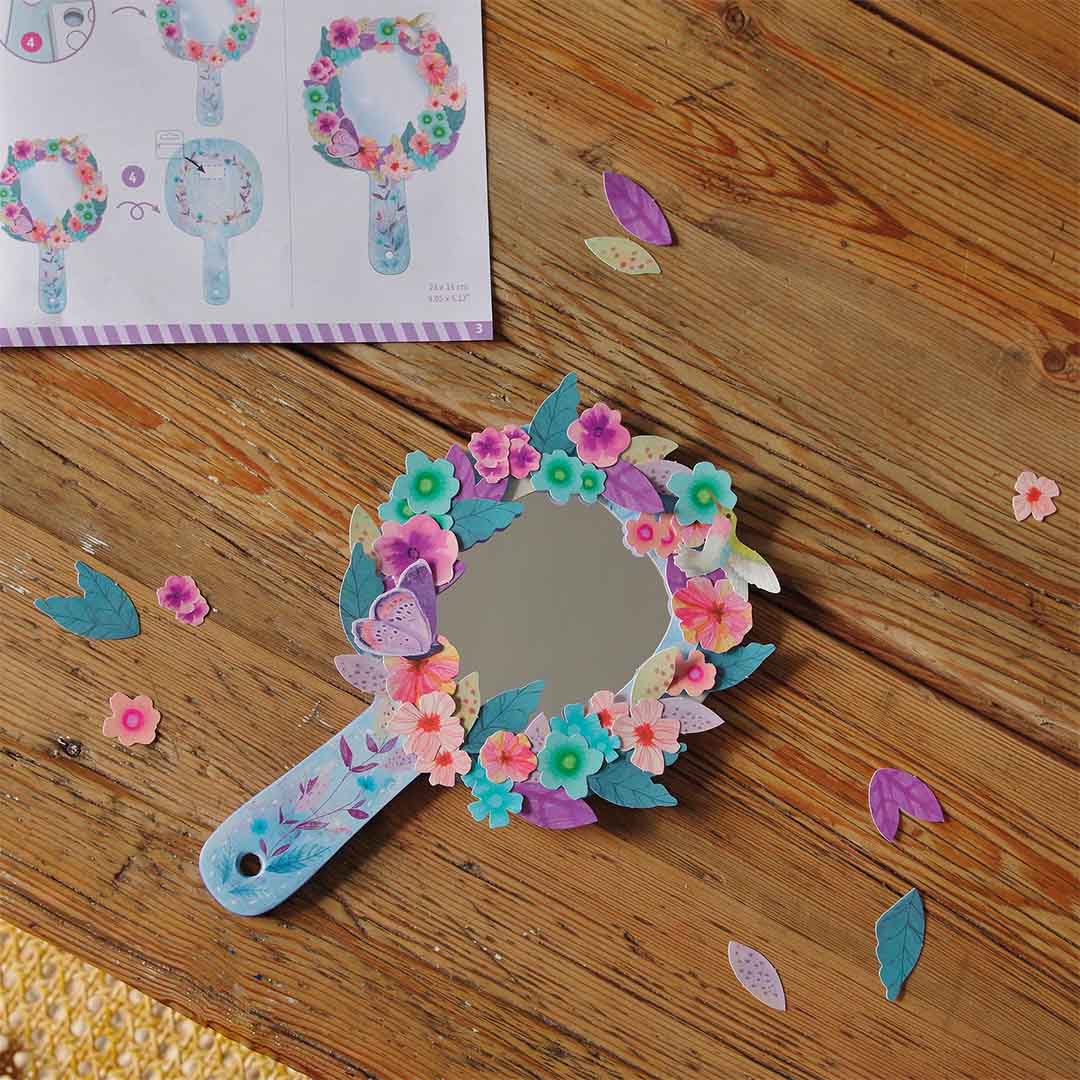Detail van een versierde spiegel van de Djeco knutselset, omringd door bloemen en vlinders op een houten tafel.