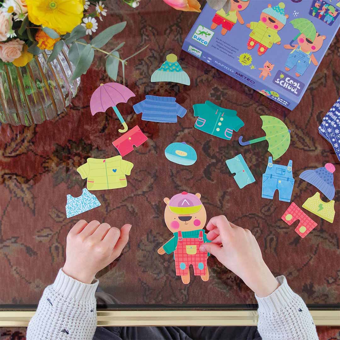 Twee kinderhanden spelen het Djeco Rapido Meteo behendigheidsspel met een beerfiguur en losse kledingstukken op een tafel.