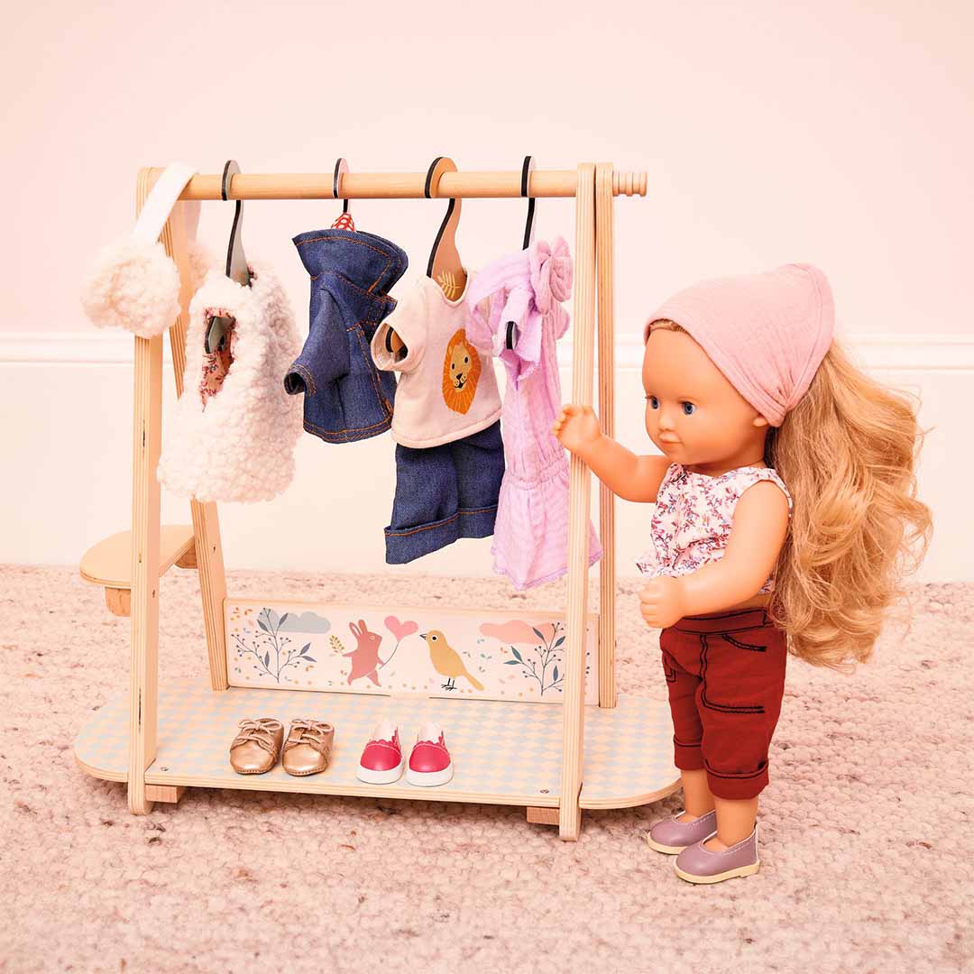 Een pop staat naast het Djeco houten poppenkledingrek met kleding en schoenen, en pakt een roze jurkje.