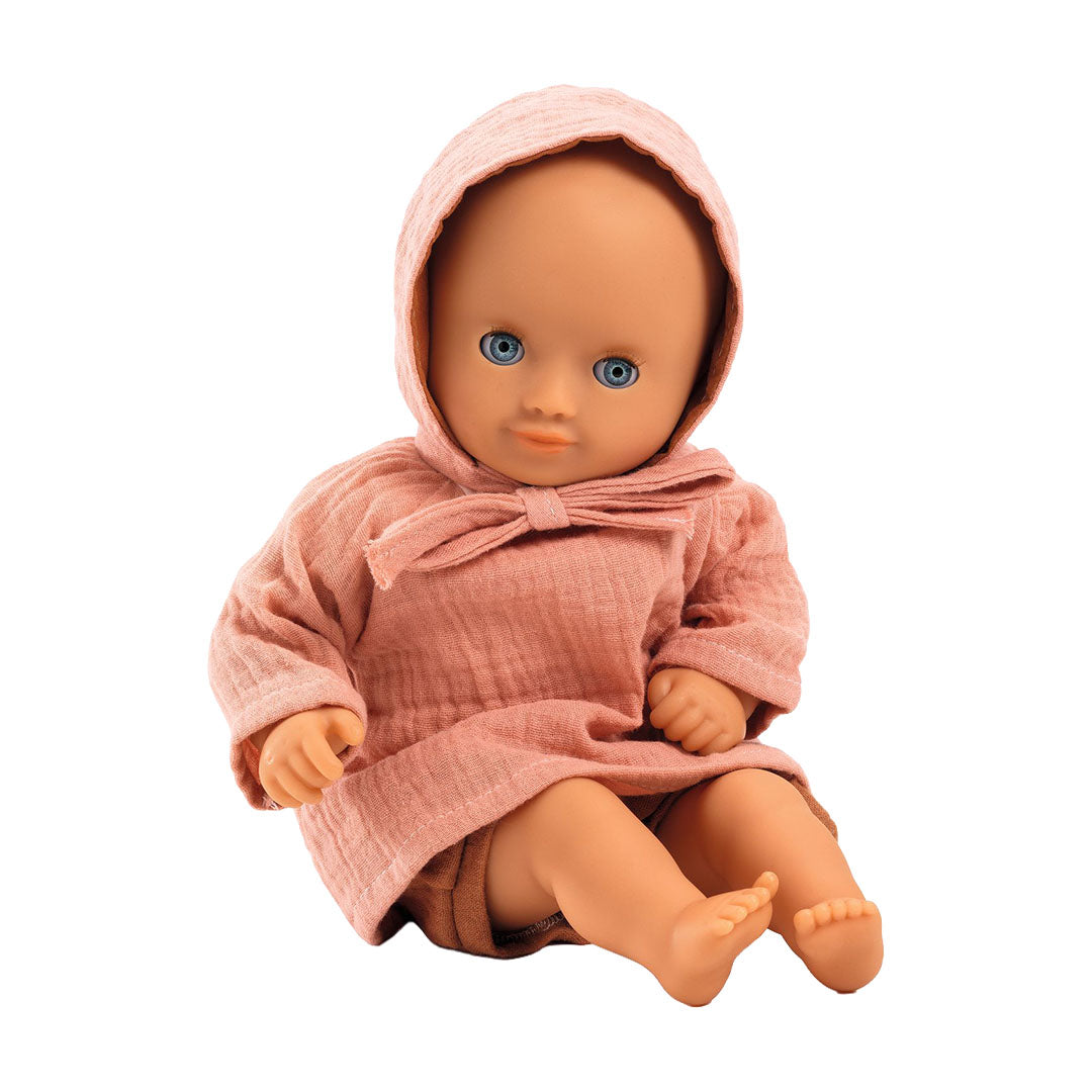 Djeco Pomea pop met blauwe ogen, zittend en gekleed in de roze top, bruine shorts en roze mutsje.