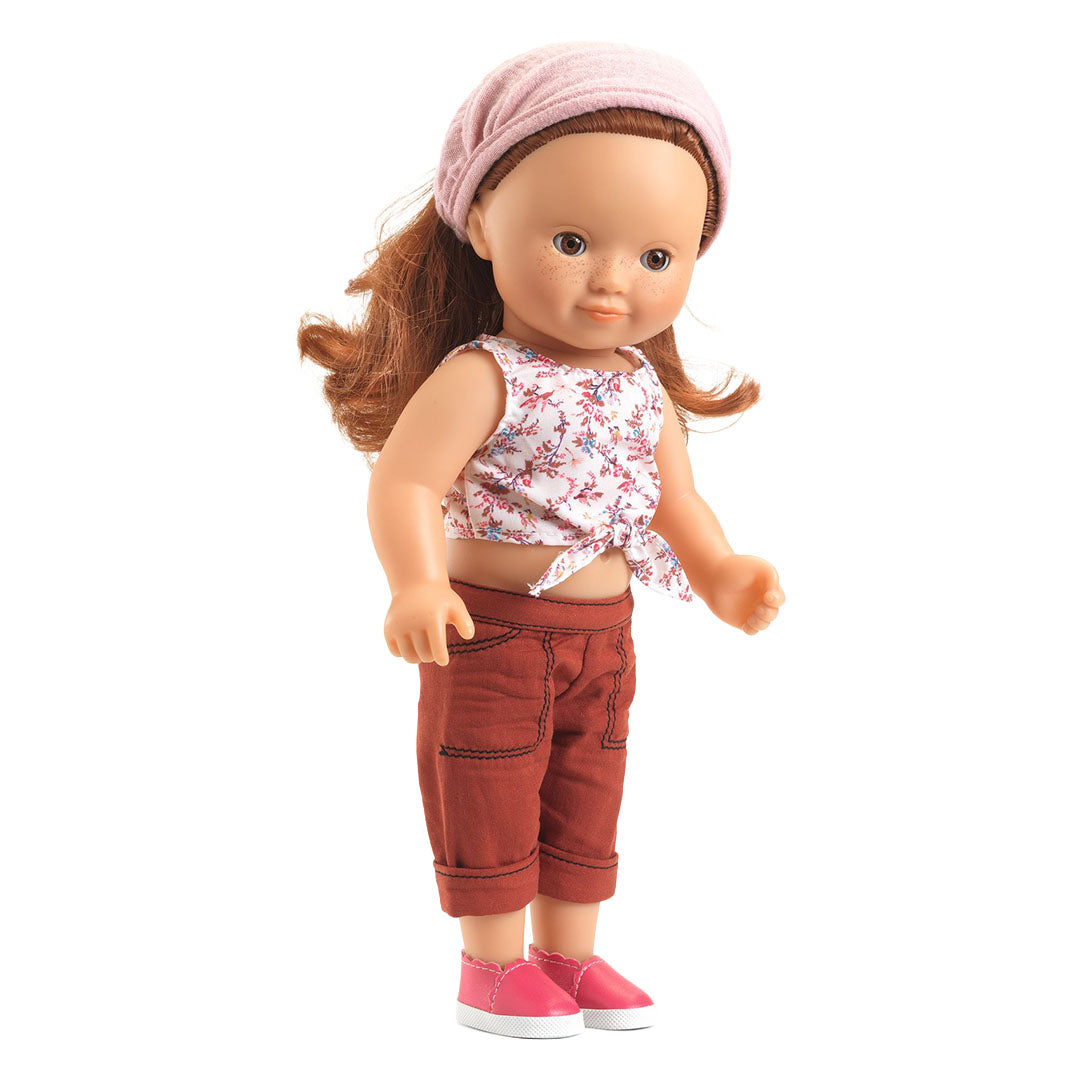 Pop met rood haar draagt de Djeco poppenkledingset met roze muts, gebloemde top en bordeauxrode broek.