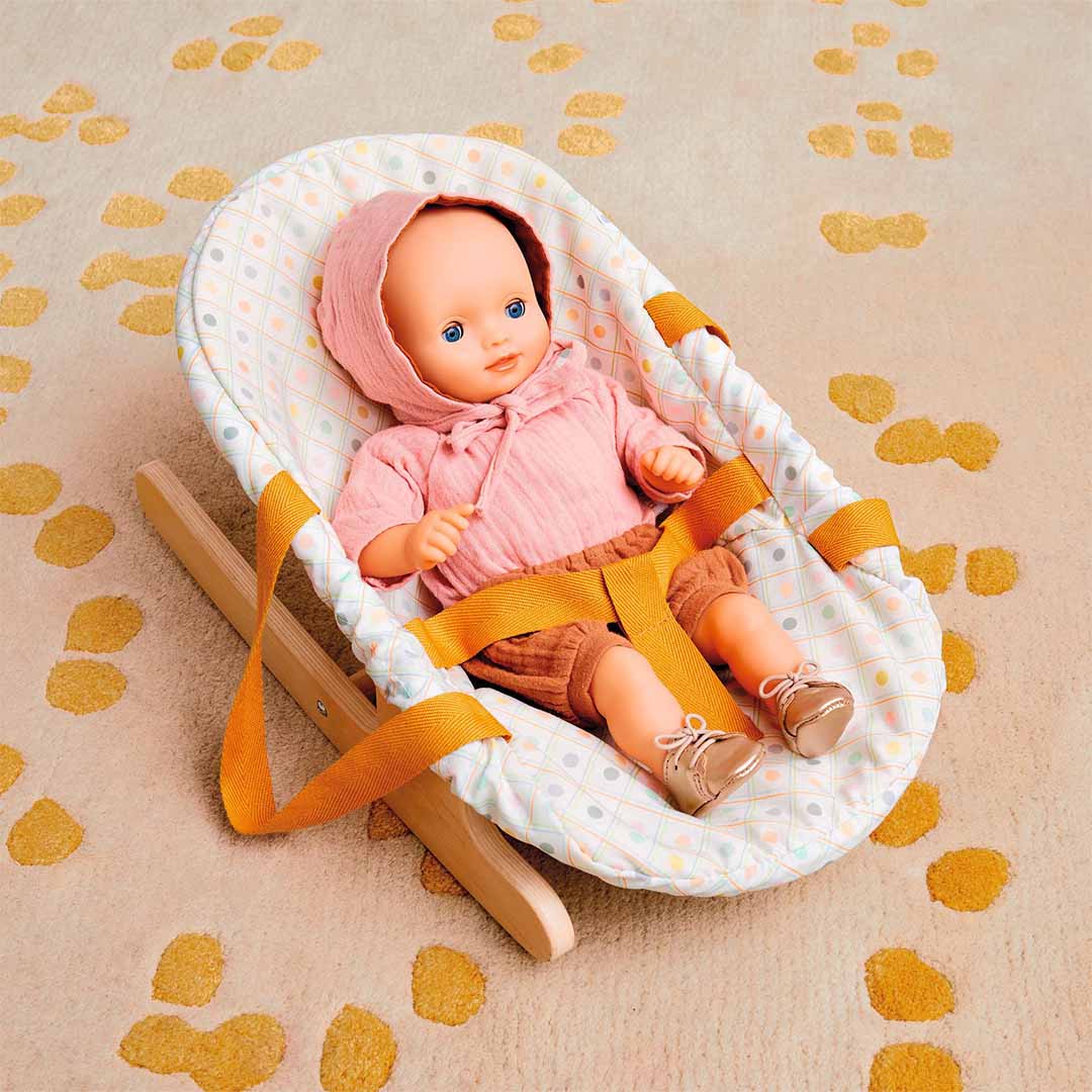 Een pop met Djeco Pomea gouden poppenschoenen en roze kleding zit in een poppenwipper op een vloerkleed.