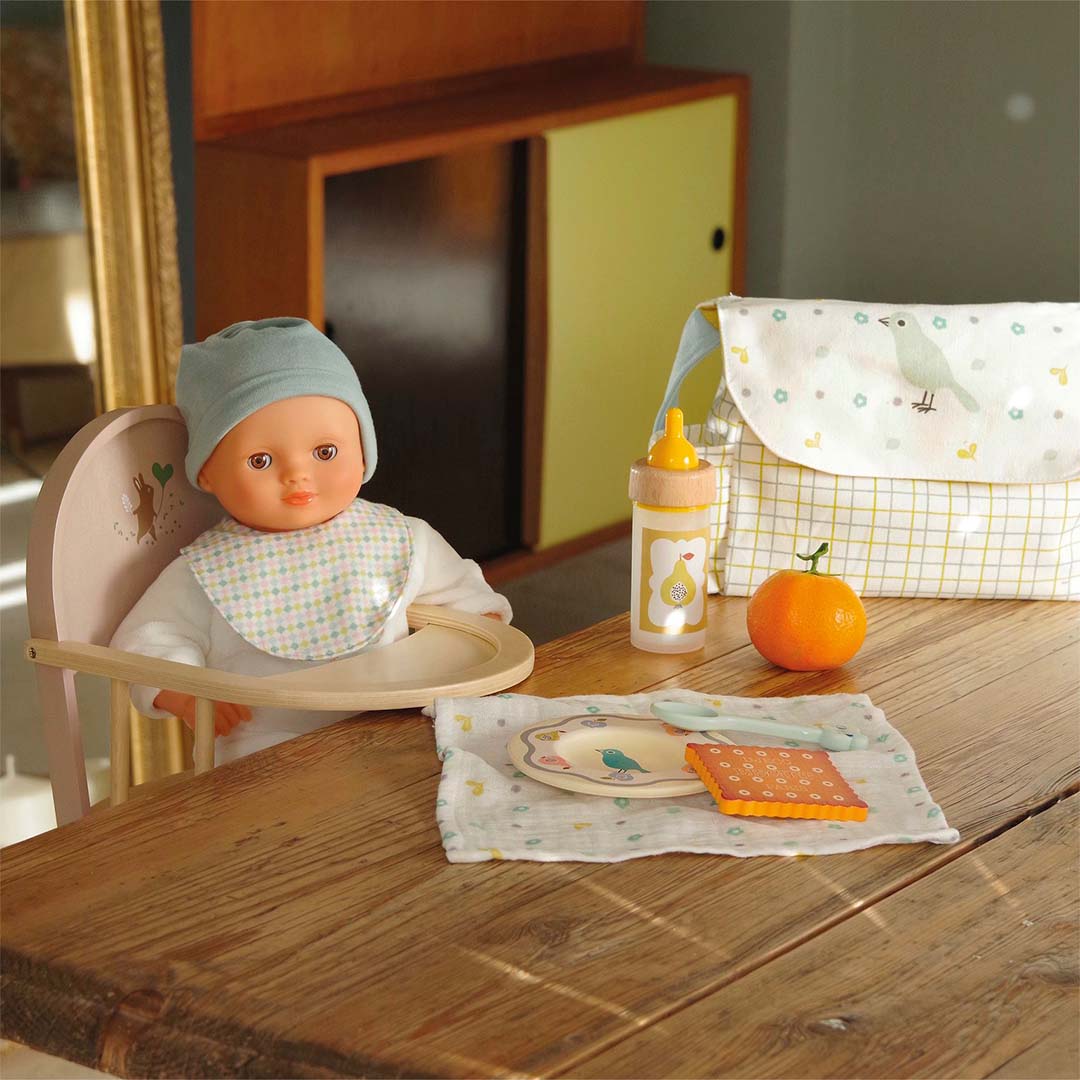 Een pop zit in een houten kinderstoel aan tafel met de Djeco Pomea poppenvoedingsset, inclusief tas en fles.