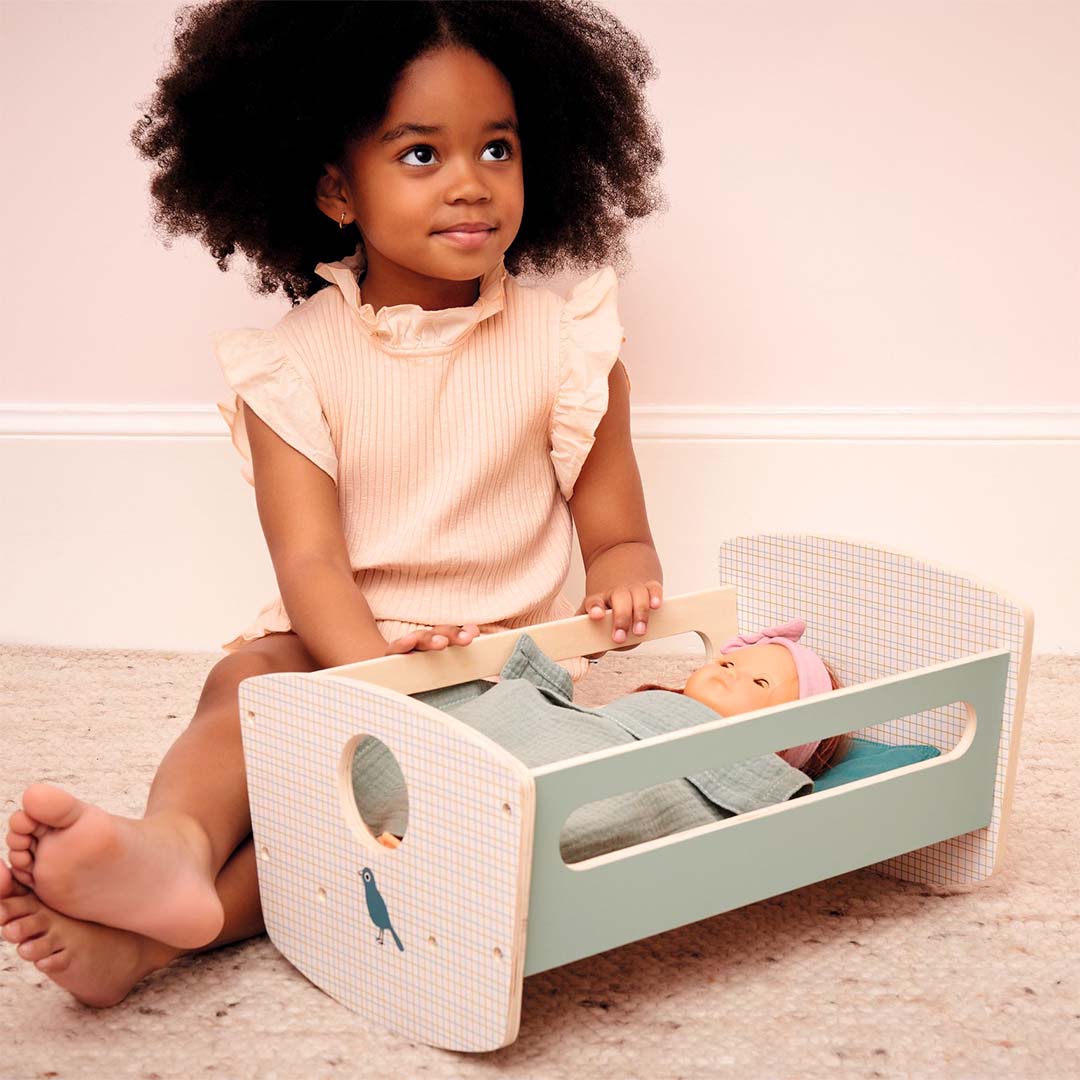 Een kind zit naast de Djeco houten poppenwieg met een pop erin.
