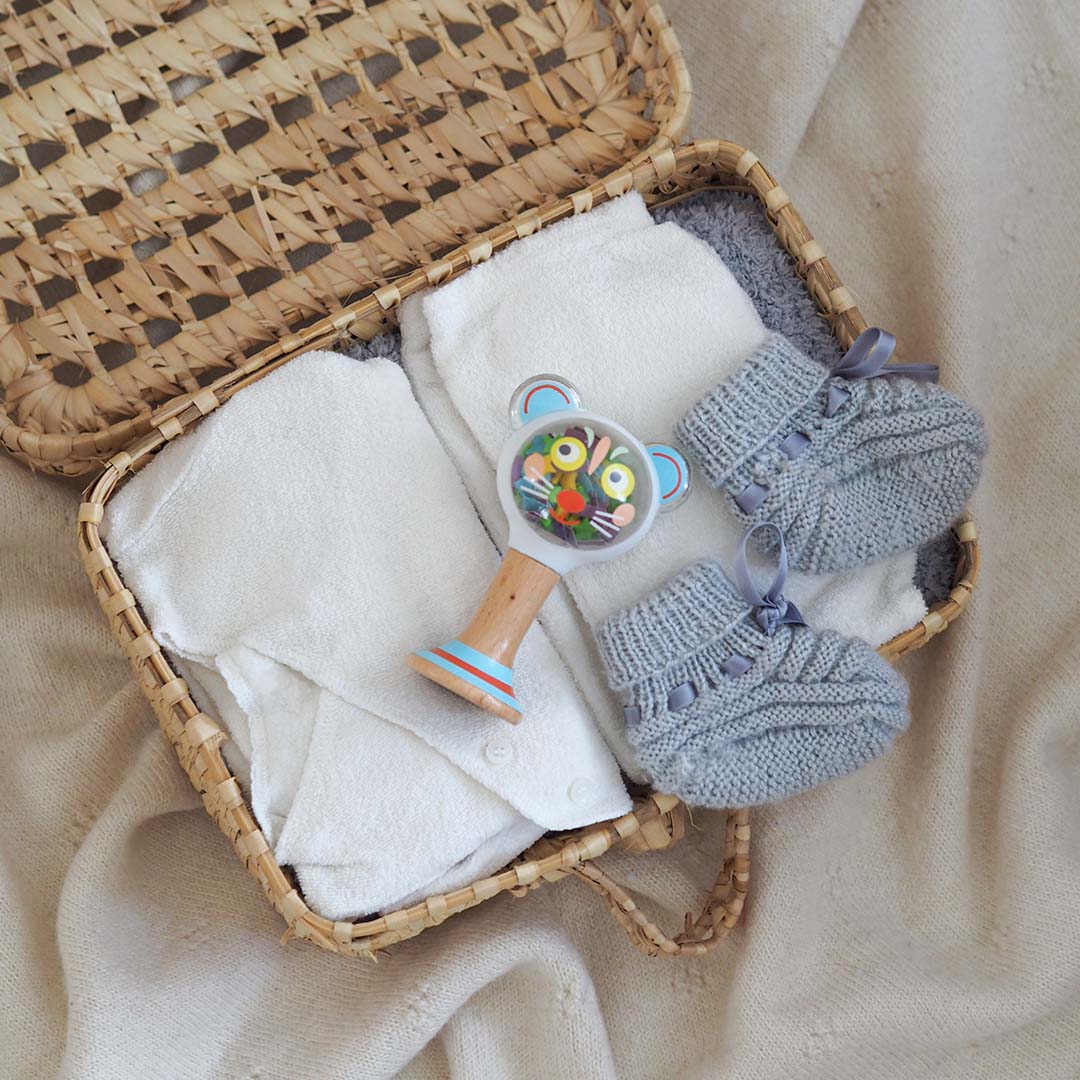 Een Djeco babyspeelgoed rammelaar ligt in een rieten mand met witte doeken en grijze gebreide babyslofjes.