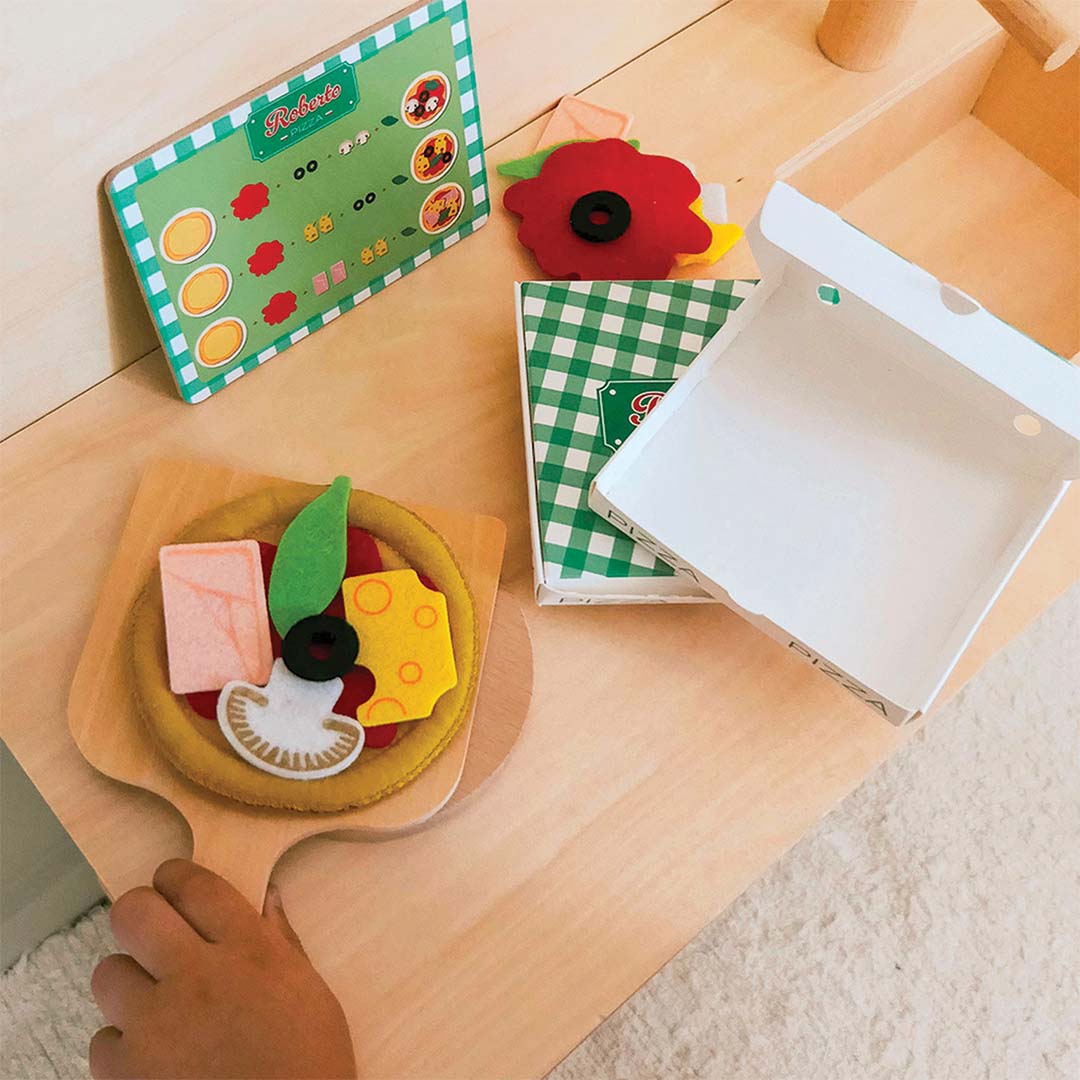Een belegde viltpizza op een houten pizzaschep, een open pizzadoos en een menukaart op een houten ondergrond.