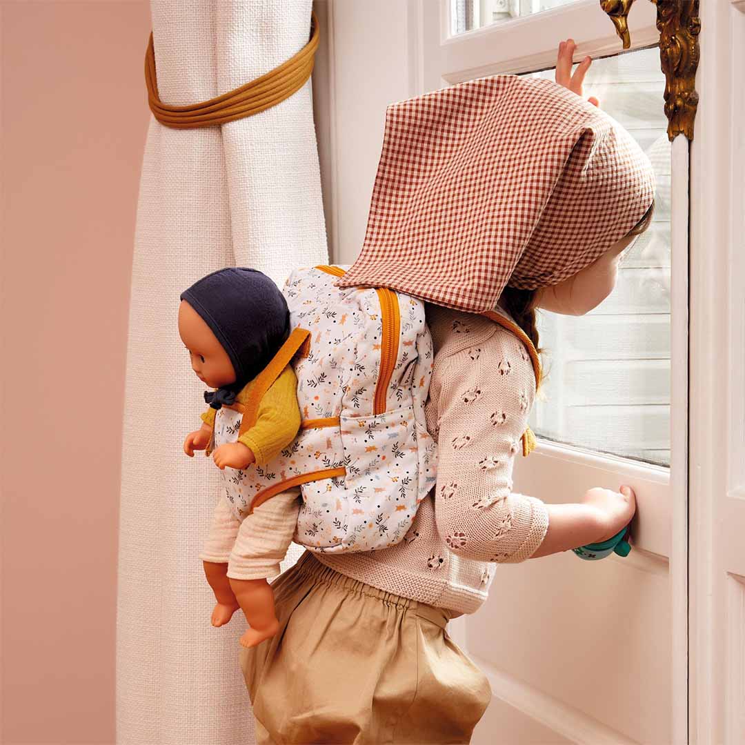 Een kind met een geruite muts draagt de Djeco poppendraagzak met pop op de rug en kijkt uit het raam.