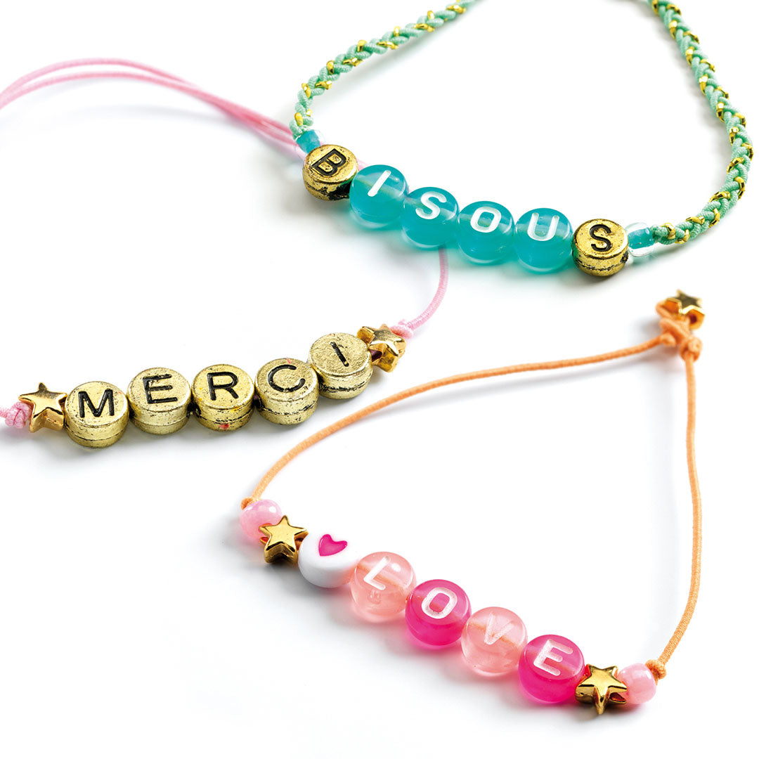 Drie gemaakte armbandjes met alfabetkralen die de woorden 'BISOU', 'MERCI' en 'LOVE' spellen.
