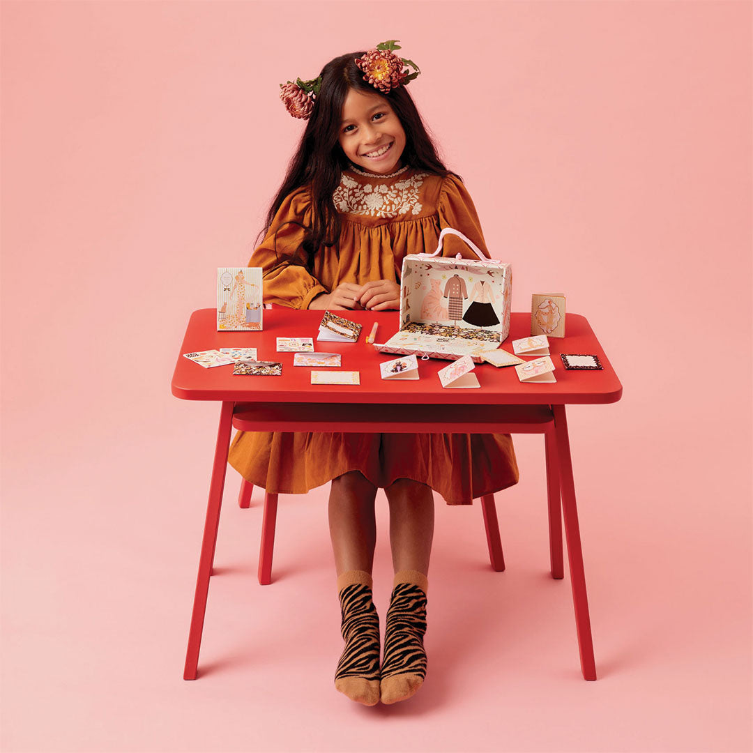 Een lachend meisje met bloemenkroon speelt aan een rode tafel met de Djeco fashion stationary hobbyset.