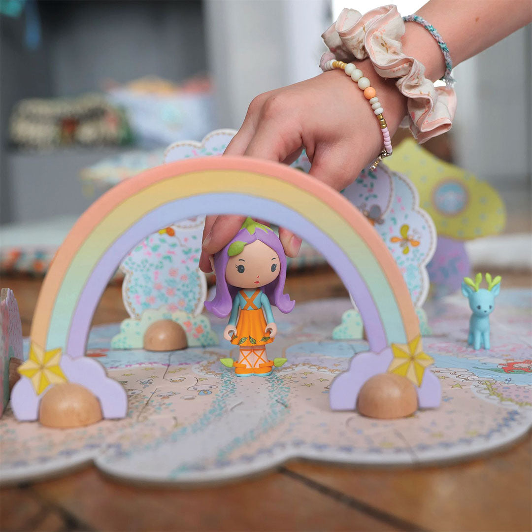 Een hand plaatst een paarse feeënpop onder een regenboogboog op de Djeco Tinyly vloerpuzzel.
