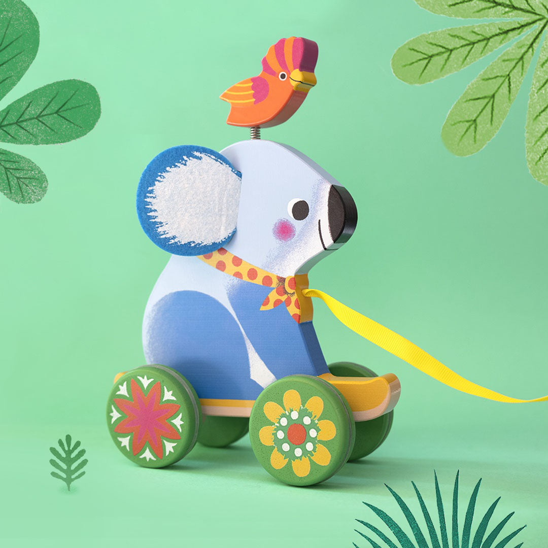 Djeco houten trekfiguur van een blauwe koala met een vogel en bloemige wielen, op een groene achtergrond met bladeren.