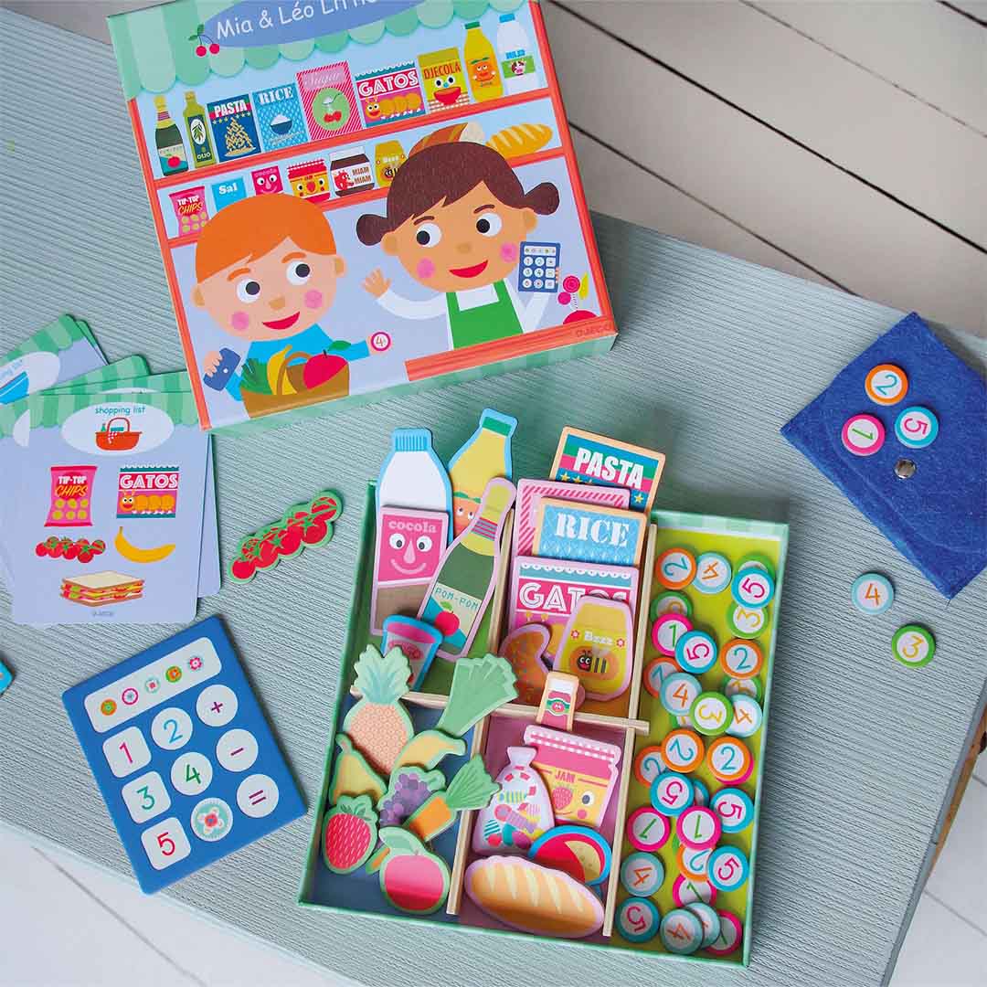 Bovenaanzicht van de Djeco Mia & Léo Little Shop speelset met houten en kartonnen speelgoedboodschappen, munten en een kassa op een blauwe ondergrond.
