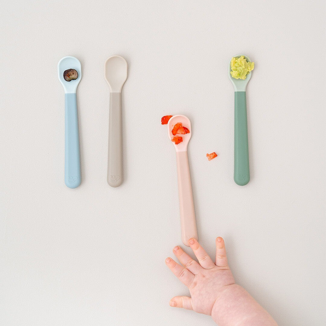 Vier Done by Deer kinderlepels in verschillende kleuren met babyvoeding erop, met een babyhandje dat naar een roze lepel reikt.