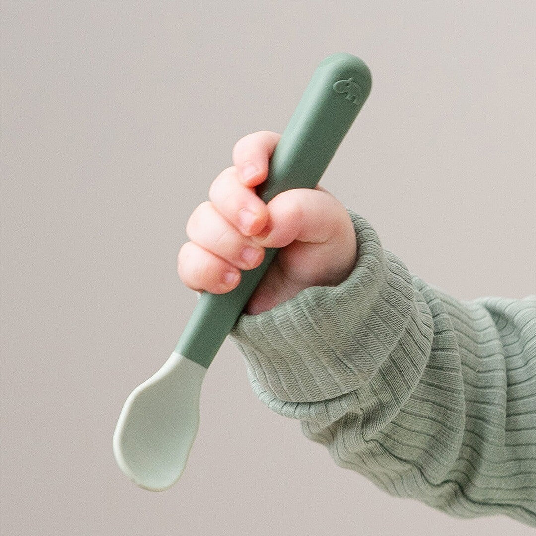 Een close-up van een babyhandje dat een groene Done by Deer kinderlepel vasthoudt.
