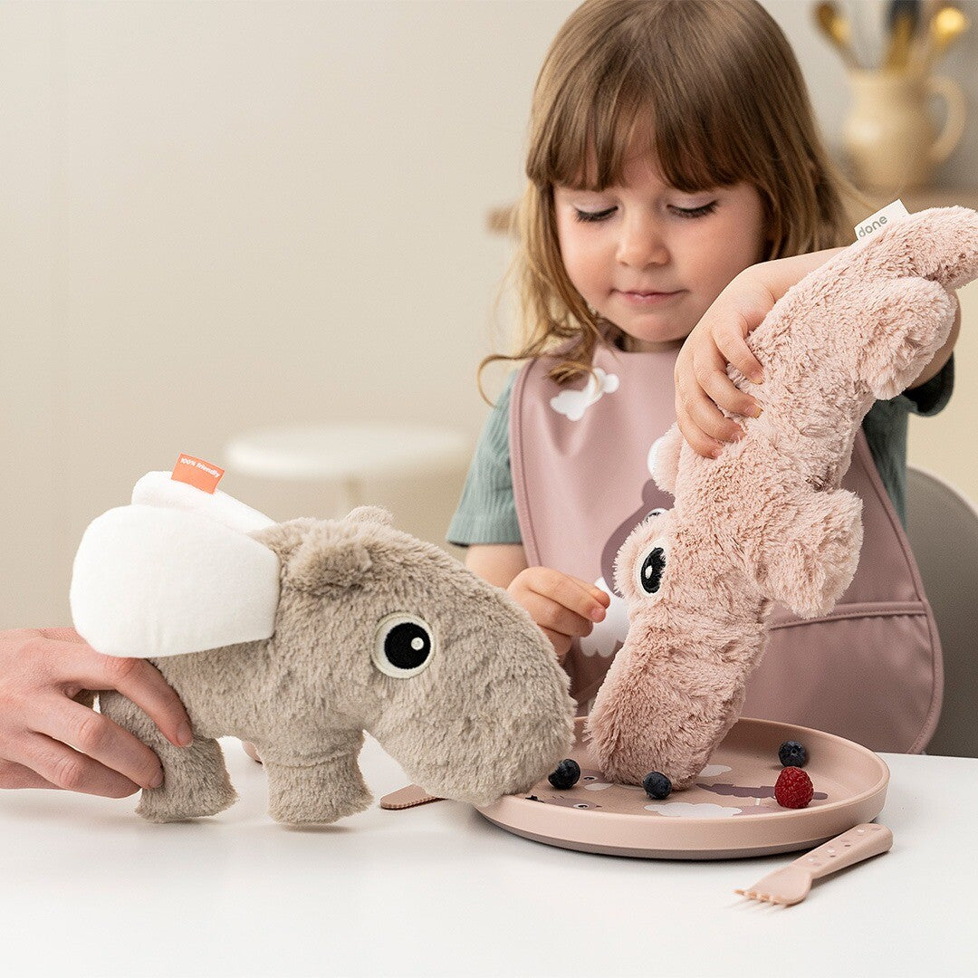 Een kind speelt aan tafel met twee Done by Deer pluchen knuffelnijlpaarden, een zandkleurige en een roze.